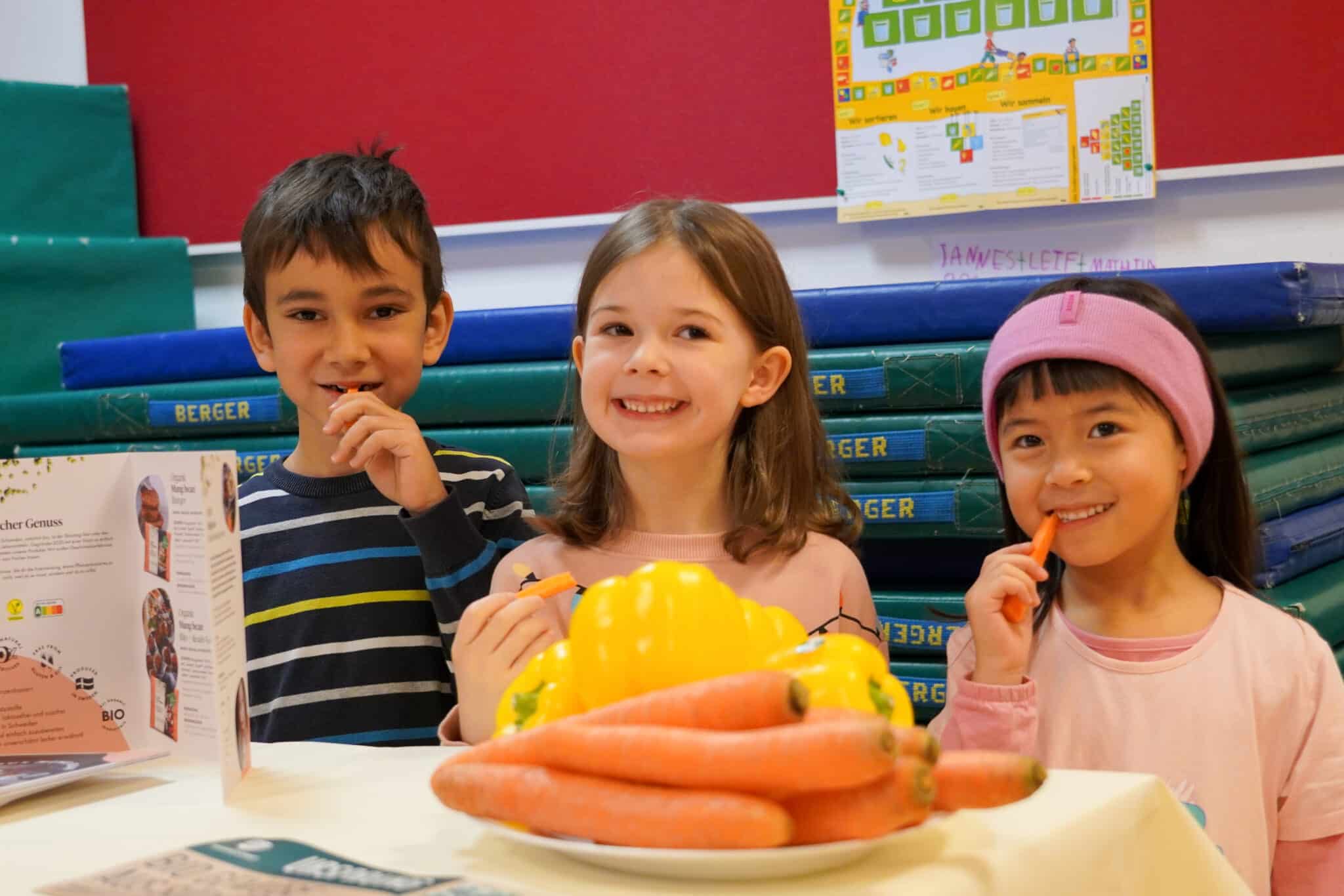 drei Kinder stehen in einer Turnhalle und essen Möhren